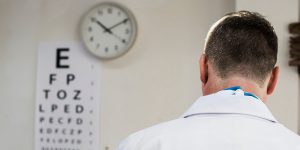 Das Bild zeigt den Rücken eines Augenarztes, im Hintergrund sieht man eine unscharfe Liste mit Buchstabenreihen