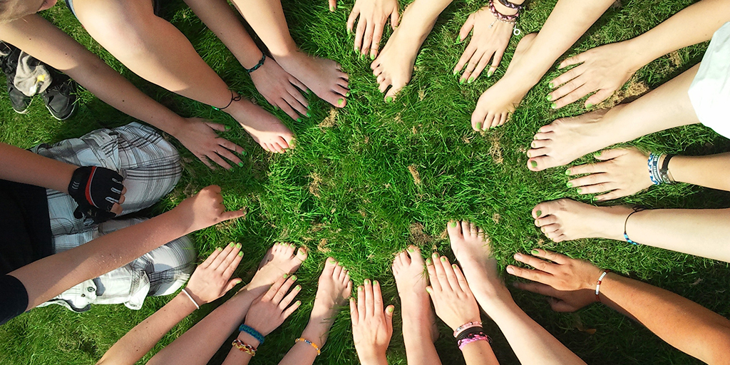 das Bild zeigt viele Hände (und ein paar nackte Füße) als Kreis auf einer Wiese - freundschaftlich & solidarisch! Foto von https://www.pexels.com/de-de/foto/beine-freundschaft-fusse-gras-53958/