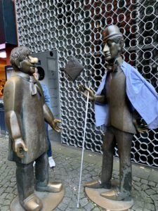 Das Bild zeigt die Kölner Eisenfiguren "". Der schielende Schäl trägt eine blaue Jacke und in seiner Hand einen weißen Langstock.