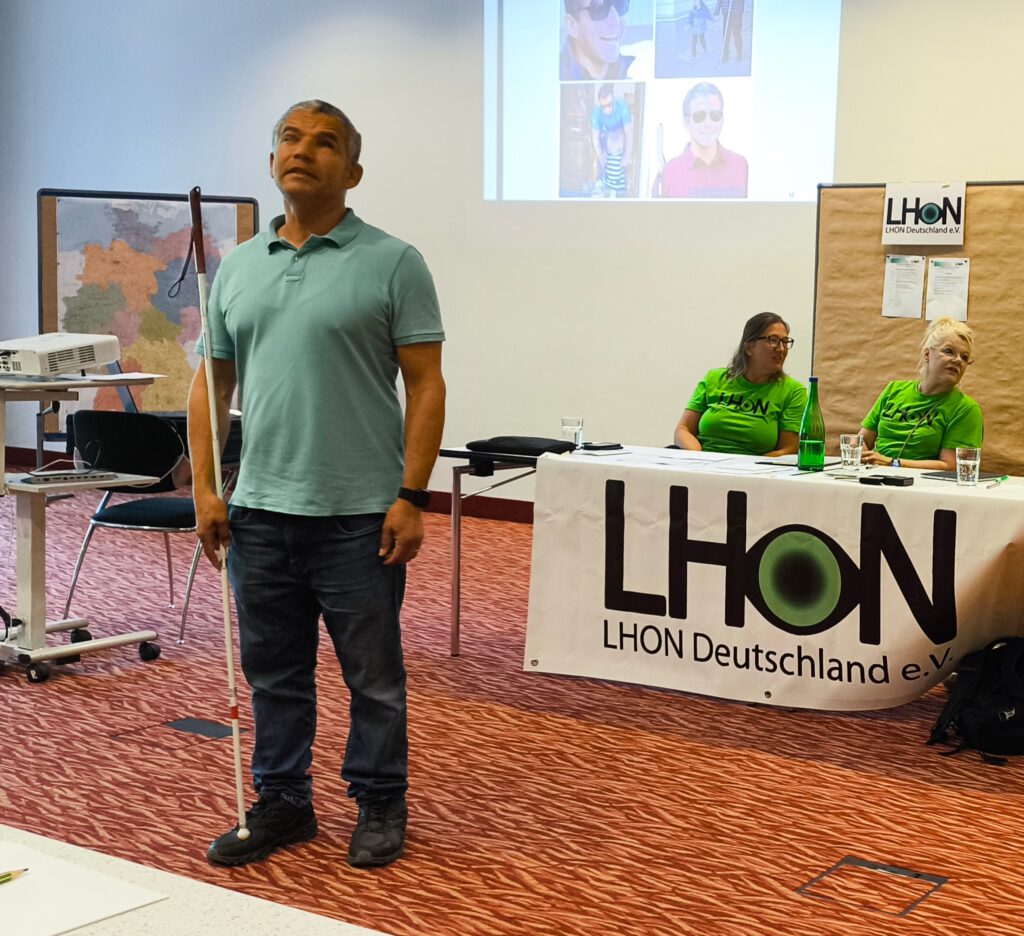 Juan steht bei seinem Vortrag im Tagungsraum vor dem Tisch des Vorstands zu den Mitglieder gewandt