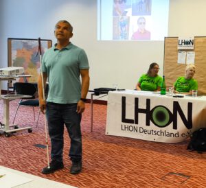 Juan steht bei seinem Vortrag im Tagungsraum vor dem Tisch des Vorstands zu den Mitglieder gewandt