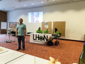 Juan steht bei seinem Vortrag im Tagungsraum vor dem Tisch des Vorstands zu den Mitglieder gewandt