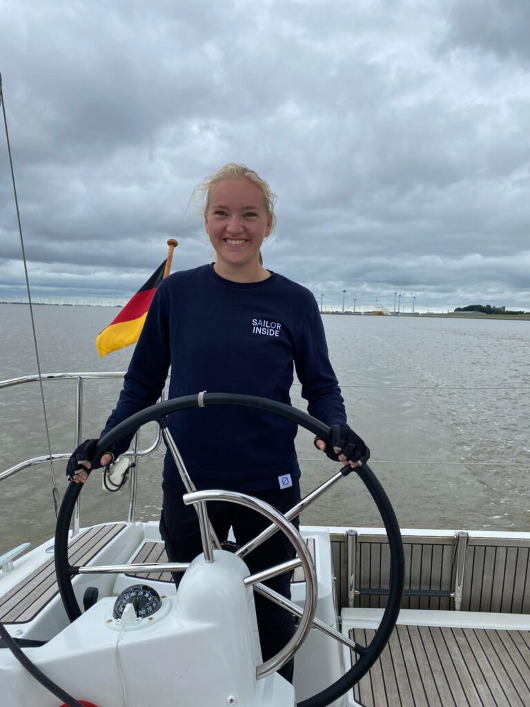 Die Jugendvertretrin Beke steht am Ruder eines Bootes mit dem Gewässer im Hintergrund