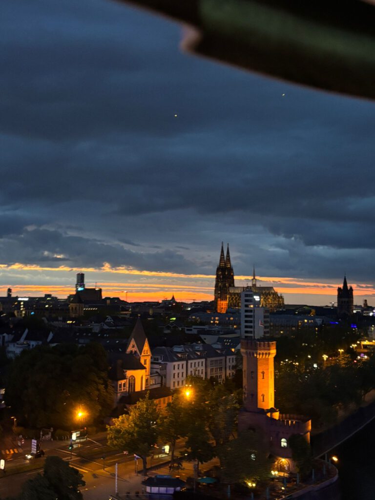 Das Bild zeigt eine stimmungsvolle nächtliche Panoramaaufnahme einer Stadtlandschaft, die aus einer erhöhten Perspektive aufgenommen wurde. Im Zentrum der Kulisse stehen historische und moderne Bauwerke vor einem farbintensiven Abendhimmel.
Der Himmel und die Lichtstimmung
Abendhimmel: Die Aufnahme entstand während der späten Dämmerung. Der obere Teil des Himmels ist von dichten, dunkelblauen bis fast schwarzen Wolkenformationen bedeckt. Zwei winzige, helle Punkte (vermutlich Sterne oder Planeten) sind im dunklen Wolkenbereich erkennbar.
Horizont: Am Horizont bricht sich das Licht der untergegangenen Sonne in einem schmalen, aber intensiv leuchtenden Band aus orangefarbenen, gelben und goldenen Tönen. Dieses glühende Licht hebt die Silhouetten der Gebäude im Hintergrund scharf ab.
Die Stadtlandschaft und markante Bauwerke
Der Kölner Dom: Etwas rechts von der Bildmitte ragt die unverkennbare, dunkel aufragende Silhouette des Kölner Doms mit seinen beiden markanten Spitzen in den farbigen Himmel. Die Basis und die filigranen Strukturen des Doms werden von der städtischen Beleuchtung sanft von unten angestrahlt.
Der Malakoffturm und das Ufer: Im rechten Vordergrund sticht ein historischer, rund gemauerter Wehrturm mit Zinnen – der Malakoffturm am Rheinufer – hervor. Er wird von warmen, gelben Scheinwerfern intensiv beleuchtet. Direkt daneben und dahinter erstrecken sich begrünte Uferbereiche und die dunkle Wasseroberfläche des Flusses am rechten unteren Bildrand.
Stadtstrukturen: Im mittleren und linken Bildbereich erstreckt sich ein dichtes Netz aus Wohn- und Geschäftshäusern mit hellen Fassaden und dunklen Dächern. Eine Kirche mit einem spitzen Turm fällt im linken mittleren Vordergrund auf. Ganz links im Hintergrund ist zudem die beleuchtete Silhouette des KölnTurms (im Mediapark) zu sehen, während rechts vom Dom der Turm von Groß St. Martin aufragt.
Straßenbeleuchtung: Die Straßen und Kreuzungen im linken Vordergrund sind durch die gelb-orangefarbenen Lichter der Straßenlaternen hell erleuchtet, was einen warmen Kontrast zu den kühlen, dunklen Tönen des restlichen Bildes bildet.