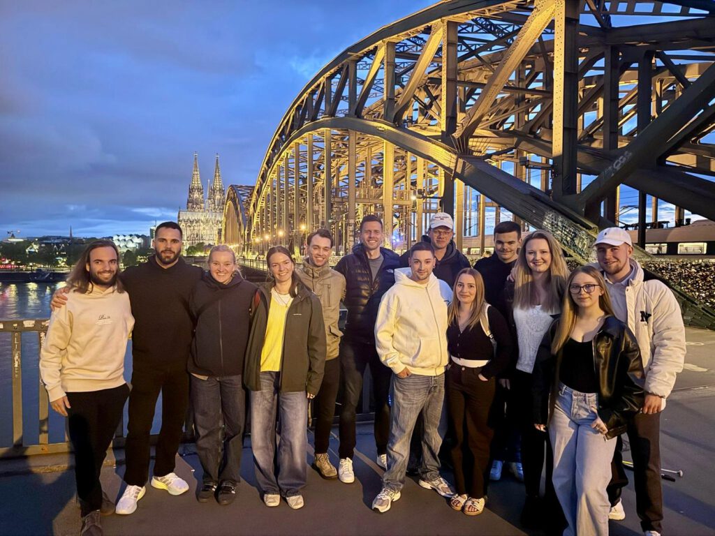 Das Bild zeigt eine Gruppe von elf jungen Erwachsenen, die sich für ein Gruppenfoto auf einer Brücke am Abend aufgestellt haben. Im Hintergrund erstreckt sich eine markante Brückenarchitektur sowie eine historische Kulisse.
Die Gruppe besteht aus sechs Männern und fünf Frauen, die nebeneinander in einer lockeren Reihe aufgereiht sind. Sie sind im Freizeit-Look gekleidet (Sweatshirts, Jacken, Jeans und Sneakers) und lächeln fast alle freundlich in die Kamera.
Linke Seite: Ganz links steht ein Mann mit längeren Haaren in einem hellen Hoodie. Daneben steht ein Mann in komplett schwarzer Kleidung, der den Arm um die Schulter einer Frau in einer dunklen Jacke gelegt hat.
Mittlerer Bereich: Es folgen eine Frau in einer dunklen Jacke mit gelbem Oberteil darunter sowie mehrere Männer – einer in einer beigen Jacke, einer in einer gesteppten Daunenjacke und einer in einem auffälligen weißen Hoodie.
Rechte Seite: Hier stehen drei Frauen und zwei Männer. Eine der Frauen trägt eine schwarze Lederjacke und eine Brille, eine andere ein weißes Spitzenoberteil. Zwei der Männer auf der rechten Seite tragen weiße Basecaps.
Hintergrund und Kulisse
Die Brücke: Die Gruppe befindet sich auf dem Fußgängerbereich einer großen Brücke. Rechts hinter ihnen ragt die massive, kunstvoll beleuchtete Stahlbogenkonstruktion der Hohenzollernbrücke empor. Das gelb-warme Licht der Brückenbeleuchtung bildet einen starken Kontrast zum tiefblauen Abendhimmel. Am Brückengeländer ganz rechts sind vage die Umrisse zahlreicher Liebesschlösser zu erkennen.
Die Stadtkulisse: Im linken Hintergrund, am anderen Ufer des Flusses (Rhein), ragt der beleuchtete Kölner Dom mit seinen markanten Zwillingstürmen in den Abendhimmel. Weiter links sind die Lichter der Kölner Altstadt und die Silhouette der Groß St. Martin Kirche zu sehen.
Der Himmel und das Wasser: Die Aufnahme wurde während der „Blauen Stunde“ gemacht. Der Himmel ist in ein tiefes Dunkelblau getaucht und leicht bewölkt. Auf der linken Seite spiegelt sich das Licht der Stadt sanft auf der Wasseroberfläche des Flusses.