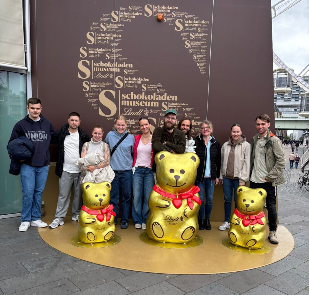 Das Bild zeigt eine Gruppe von zehn Personen, die sich für ein Erinnerungsfoto vor einer markanten Markenwand im Außenbereich aufgestellt haben.
Die Personengruppe und die Goldbären
Die Gruppe hat sich in einer Reihe auf einer halbkreisförmigen, goldfarbenen Bodenplatte formiert. Vor ihnen sind drei glänzende, goldene Bärenskulpturen (Lindt-Teddy) mit roten Halsschleifen platziert.
Linker Bereich: Ganz links steht ein junger Mann in einem dunklen „Union“-Hoodie und blauen Jeans, der eine Jacke über dem Arm hält. Daneben stehen ein Mann in einer offenen schwarzen Jacke über einem weißen T-Shirt sowie eine Frau in einem hellen, ärmellosen Oberteil, die eine weiße Jacke vor sich hält.
Mittlerer Bereich: Es folgen zwei Frauen in Freizeitkleidung (eine im hellblauen Kapuzenpullover, eine im rosafarbenen Top mit roter Strickjacke). In der Mitte lehnt sich ein Mann mit Bart, Brille und grüner Cap lässig auf den großen, zentralen Goldbären. Direkt hinter ihm schaut ein weiterer Mann mit längeren Haaren hervor, auf dessen Schulter ein winziger weißer Plüschbär sitzt.
Rechter Bereich: Rechts vom großen Bären steht eine Frau mit Brille in einer dunklen Steppjacke, gefolgt von einer jüngeren Frau in einer hellen Bomberjacke. Ganz rechts außen schließt ein junger Mann in einer olivgrünen Jacke und dunkler Hose die Reihe ab.
Hintergrund und Kulisse
Die Markenwand: Direkt hinter der Gruppe befindet sich eine große, dunkelbraune Wand. Darauf ist mit goldener Schrift eine Wortwolke in Form eines großen Kakaomotivs oder einer Bohne gestaltet. Der wiederkehrende Text lautet „schokoladen museum Lindt“ in Kombination mit einem großen, geschwungenen „S“.
Die Umgebung: Auf der rechten Bildseite öffnet sich der Blick auf eine gepflasterte Promenade, auf der vereinzelte Fußgänger unterwegs sind. Ganz im Hintergrund ist die weiße Stahlkonstruktion eines Riesenrads sowie moderne Wohnbehandlung (die Kölner Kranhäuser) unter einem bewölkten Himmel zu erkennen.