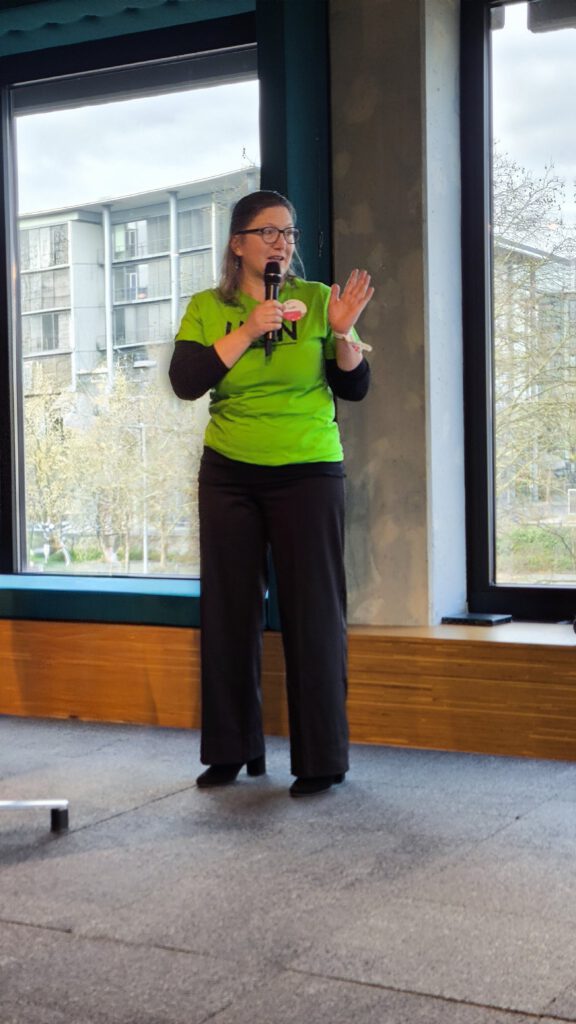 Das Bild zeigt eine Frau, die in einem hellen, modern gestalteten Innenraum einen Vortrag hält.
Die Belichtung ist natürlich und hell, da das Licht von hinten und den Seiten einfällt. Die Szene vermittelt eine professionelle, engagierte und kommunikative Atmosphäre während einer Veranstaltung.
Sie trägt ein auffälliges, leuchtend grünes T-Shirt mit einem schwarzen Aufdruck auf der Brust (teilweise verdeckt, das LHON-Logo). Darunter trägt sie ein langärmeliges, schwarzes Oberteil. Kombiniert ist das Outfit mit einer weiten, schwarzen Hose und dunklen Schuhen mit leichtem Absatz. An ihrer linken Brust ist ein kleines, rundes Namensschild angebracht. Sie trägt zudem eine Brille mit dunklem Gestell.