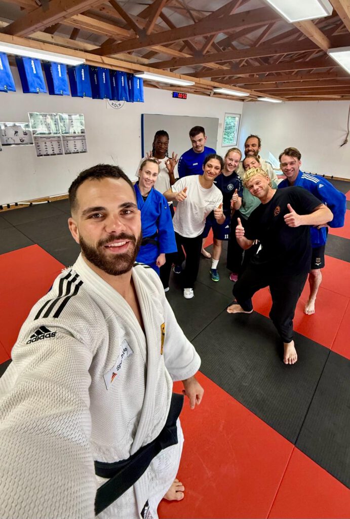 Das Bild zeigt eine Gruppe von zehn sportlich gekleideten Personen in einer Trainingshalle (Dojo), die sich für ein dynamisches Selfie aufgestellt haben.
Die Personen im Vorder- und Hintergrund
Der Selfie-Fotograf (Vordergrund): Im linken Vordergrund nimmt Lennart, ein Mann mit Bart, das Selfie auf. Er trägt einen traditionellen weißen Judo-Anzug (Judogi) der Marke Adidas mit den charakteristischen drei schwarzen Streifen auf der Schulter sowie einen schwarzen Gürtel. Auf seiner linken Brust ist ein Aufnäher der „Sporthilfe“ mit der deutschen Flagge zu sehen. Er blickt lächelnd direkt in die Kamera.
Die Gruppe (Hintergrund): Hinter ihm hat sich die restliche Gruppe von neun Personen im Raum verteilt. Sie barfuß auf den Matten, wirken ausgelassen und gut gelaunt:
Direkt hinter dem Fotografen steht eine Frau in einem leuchtend blauen Judo-Anzug, ebenfalls mit einem schwarzen Gürtel.
In der Mitte posiert eine Frau in einem weißen T-Shirt und dunkler Hose mit zwei nach oben gestreckten Daumen.
Rechts im Vordergrund sticht ein Mann mit blonden Haaren in komplett schwarzer Sportkleidung hervor, der sich dynamisch vorbeugt und ebenfalls beide Daumen anhebt.
Dahinter stehen weitere Gruppenmitglieder, teils in blauer Sportkleidung (darunter ein Mann in einem blauen Judo-Anzug ganz rechts), teils in Freizeit-T-Shirts. Eine Frau im hinteren linken Bereich formt mit beiden Händen ein Zeichen.
