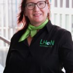 Das Foto zeigt Sandra Findeisen, eine Frau mit auffällig roten Haaren und einer Brille. Sie trägt das schwarze "LHON Deutschland e.V."-Hemd und ein grünes Halstuch. Ihr Lächeln ist herzlich und authentisch. Der Hintergrund zeigt eine moderne Glasfassade. Das Licht ist etwas kühl, aber dennoch angenehm.