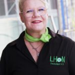 Dieses Portrait zeigt Birte Preuß, eine Frau mit platinblonden, hochgesteckten Haaren und einer Brille mit Metallgestell. Sie trägt ein schwarzes Hemd mit dem grünen "LHON Deutschland e.V."-Logo und ein passendes grünes Halstuch. Sie lächelt freundlich in die Kamera. Der Hintergrund zeigt einen unscharfen Gebäudeteil und etwas Grünfläche. Die Beleuchtung ist hell und freundlich.