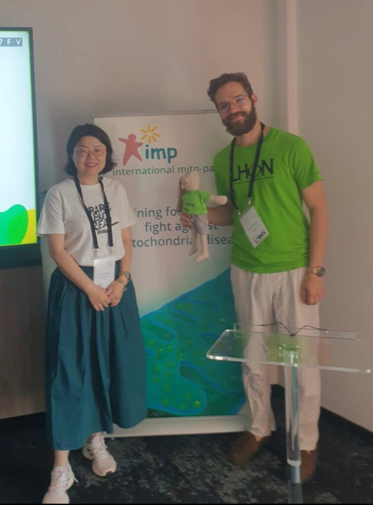 Dieses Foto zeigt die chinesische Vertreterin (links) und Tom Schuster, einen bärtigen Mann (rechts), die in einem Innenraum vor einem Werbe-Roll-up-Banner stehen. Die Frau trägt ein weißes T-Shirt mit der schwarzen Aufschrift „RARE IS BUT REAL“ in Fettschrift und eine blaue Hose. Sie lächelt in die Kamera und hat ihre Hände vor sich verschränkt. Der Mann trägt ein neongrünes T-Shirt mit der schwarzen Aufschrift „LHON“ und helle Hosen. Er hält in seiner rechten Hand eine kleine Puppe mit einem passenden neongrünen T-Shirt, auf dem ebenfalls „LHON“ steht. Beide haben Ausweise an Schlüsselbändern um den Hals.
Das Roll-up-Banner hinter ihnen ist weiß und grün und zeigt das Logo der „International Mito-Patients“ (IMP) sowie die Aufschrift „Joing forces in the fight against Mitochondrial disease“. Ein Teil des Textes auf dem Banner ist durch die Personen verdeckt. Links im Hintergrund ist ein Bildschirm mit einer grünen und weißen Grafik zu sehen. Davor steht ein durchsichtiges Rednerpult. Der Boden ist mit einem grauen Teppich ausgelegt.