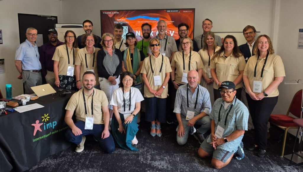 Ein Gruppenfoto, das 21 Personen in einem Konferenz- oder Tagungsraum zeigt. Die Gruppe steht und hockt vor einem Tisch und einem großen Bildschirm. Die Stimmung wirkt positiv und professionell.
Komposition und Personen:
Das Bild ist eine Weitwinkelaufnahme, die die Gruppe in drei Reihen zeigt: Eine vordere Reihe, die hockt, und zwei stehende Reihen dahinter.
Vordergrund (Hockende Reihe):
Vier Personen hocken am Boden. Von links nach rechts:
Ein Mann in einem beigen Poloshirt, dunkelblauer Hose und hellen Schuhen. Er trägt ein Namensschild an einem Umhängeband.
Eine Frau in einem weißen T-Shirt mit der Aufschrift „RARE BUT REAL“, einer weiten blauen Hose und weißen Turnschuhen.
Ein Mann in einem blassblauen, gemusterten Kurzarmhemd und hellgrauer Hose. Er trägt ebenfalls ein Namensschild.
Ein Mann in einem hellblauen, karierten Kurzarmhemd, grauen Shorts und einer dunkelgrünen Schirmmütze mit dem weißen Logo „POLG“. Er trägt ein Namensschild und lächelt in die Kamera.
Mittlere Reihe (Stehend, vorderer Teil):
Sieben Personen stehen direkt hinter den Hockenden. Fünf von ihnen tragen identische, hellbeige Poloshirts mit einem kleinen grünen Logo auf der linken Brustseite und Namensschilder an Umhängebändern.
Von links nach rechts:
Ein Mann im beigen Poloshirt und dunkler Hose.
Eine Frau im beigen Poloshirt und einer dunklen, gemusterten Hose/Rock.
Eine Frau im beigen Poloshirt und weißer Hose.
Die zentrale Person der Gruppe: Eine Frau mit kurzen, hellen Haaren, die eine dunkle Sonnenbrille trägt, ein beiges Poloshirt, eine dunkle Hose und offene Schuhe. Sie hält ihre Hände vor sich verschränkt und trägt ein Namensschild.
Eine Frau im beigen Poloshirt und dunkler Hose.
Eine Frau im beigen Poloshirt und dunkler Hose.
Eine Frau im beigen Poloshirt und dunkler Hose, am rechten Rand dieser Reihe.
Hintere Reihe (Stehend):
Zehn Personen stehen in der hintersten Reihe. Ihre Kleidung ist vielfältiger.
Von links nach rechts:
Ein Mann in einem hellblauen Button-Down-Hemd und grauer Hose.
Ein Mann in einem dunkelvioletten Hemd und einer schwarzen Schirmmütze mit dem Logo „POLG“.
Ein Mann in einem dunklen Hemd, der teilweise von der Person davor verdeckt wird.
Ein Mann in einem hellgrauen Hemd.
Ein Mann, fast ganz verdeckt.
Ein Mann in einem grünen T-Shirt mit dem Logo „LHON“ und dunkler Hose.
Ein Mann in einem grauen Hemd und dunkler Hose.
Ein Mann, teilweise verdeckt, in einem dunklen Oberteil.
Ein Mann in einem grauen Hemd und dunkler Hose.
Ein Mann in einem blauen Anzug, am rechten Rand der hintersten Reihe.
Umgebung und Hintergrund:
Linker Bildrand:
Ein Tisch ist mit einem schwarzen Tuch bedeckt, das als Werbebanner dient. Darauf ist das bunte Logo „IMP“ zu sehen, bestehend aus einem stilisierten pinkfarbenen Stern-Mensch-Symbol und dem Text „international mito patients“. Auf dem Tisch stehen verschiedene Gegenstände: Eine türkisfarbene Wasserflasche, eine Brille in einem Etui, ein aufgeklappter Laptop (teilweise sichtbar), Stifte, Becher und Papiere.
Mitte/Hintergrund:
Ein großer Flachbildfernseher ist an der weißen Wand befestigt. Er zeigt eine bunte Grafik, die wie eine Berglandschaft aussieht, und einige Texte, die jedoch zu klein sind, um deutlich gelesen zu werden. Oben rechts auf dem Bildschirm stehen Zahlen und ein Code: „CODE W3AWWK“ und „2026-06-16 11:27“. Über dem Bildschirm ist links ein weißer Projektor an der Decke sichtbar.
Rechter Bildrand:
Am rechten Rand stehen einige rote Stühle, die an der Wand lehnen. Der Boden ist mit einem dunklen, gemusterten Teppich ausgelegt. Die Wände sind weiß gestrichen.
Atmosphäre und Stil:
Die Beleuchtung ist natürlich, als ob das Foto am helllichten Tag aufgenommen wurde. Alle Personen blicken in die Kamera, viele lächeln. Das Foto wirkt wie ein spontaner Schnappschuss, um eine Gruppe von Teilnehmern einer Veranstaltung oder Konferenz festzuhalten. Die Namensschilder und die einheitlichen Poloshirts lassen auf eine offizielle Tagung oder ein Treffen schließen.