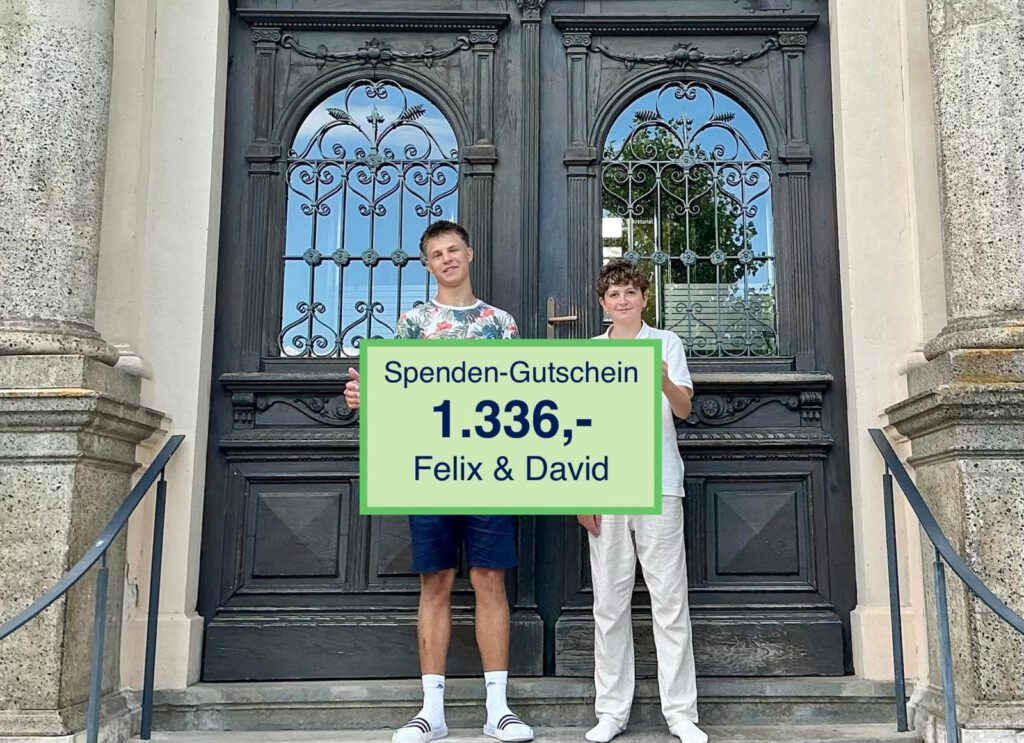 Felix und David stehen vor einer verzierten, dunklen Holzdoppeltür mit schmiedeeisernen Fenstern. Sie halten ein großes grünes Schild mit der Aufschrift „Spenden-Gutschein 1.336,- Felix & David“