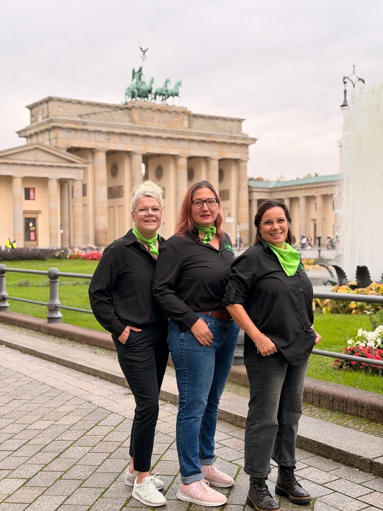 Das Foto zeigt von links nach rechts Birte, Sandra und Francesca, die nebeneinander auf einem gepflasterten Weg stehen und lächelnd in die Kamera blicken. Alle drei tragen einheitliche Kleidung: schwarze Blusen beziehungsweise Hemden mit dem grünen Vereinslogo auf der linken Brustseite sowie auffällige, leuchtend grüne Halstücher. Dazu tragen sie dunkle Stoffhosen oder Jeans und flache, sportliche Schuhe. Hinter den Frauen befindet sich ein niedriges Metallgeländer mit einer Rasenfläche und Blumen. Im Hintergrund erhebt sich das Brandenburger Tor unter einem hellen, bewölkten Himmel. Am rechten Bildrand ist zudem die hohe, weiße Wasserfontäne eines Springbrunnens angeschnitten.